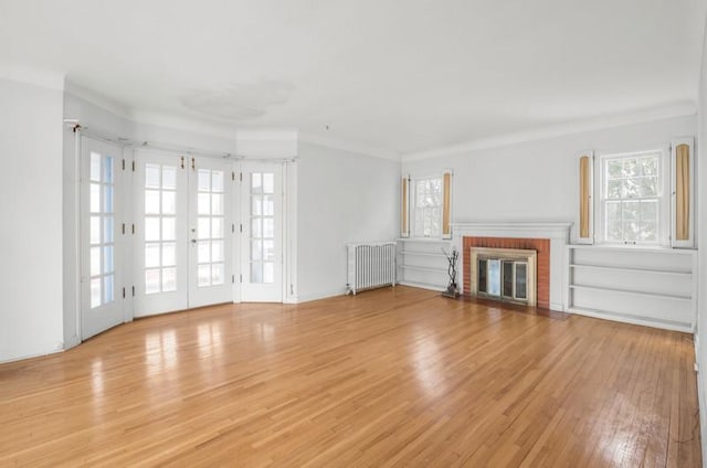 unfurnished living room featuring french doors, crown molding, radiator heating unit, a brick fireplace, and light wood finished floors