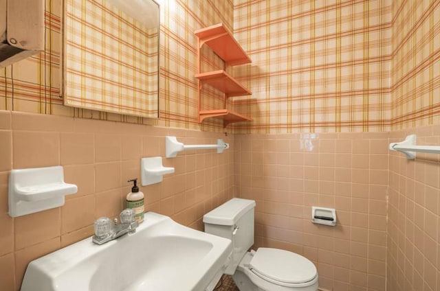 bathroom with wallpapered walls, toilet, wainscoting, tile walls, and a sink