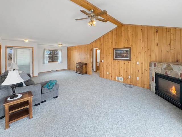 carpeted living room with a ceiling fan, vaulted ceiling with beams, a fireplace, wood walls, and visible vents