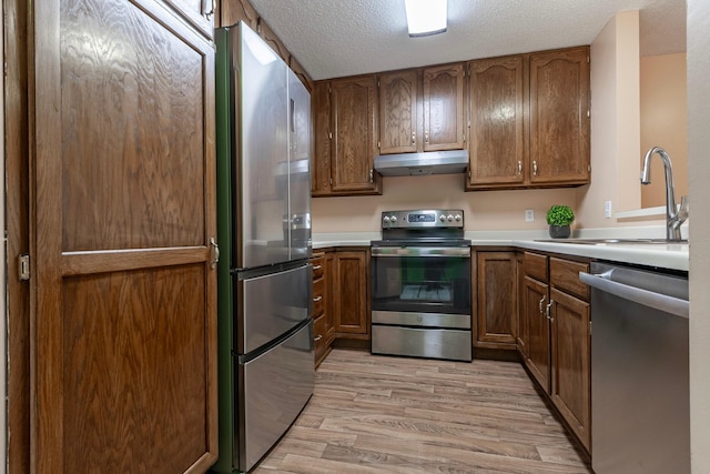 kitchen with brown cabinets, under cabinet range hood, a sink, light wood-type flooring, and appliances with stainless steel finishes
