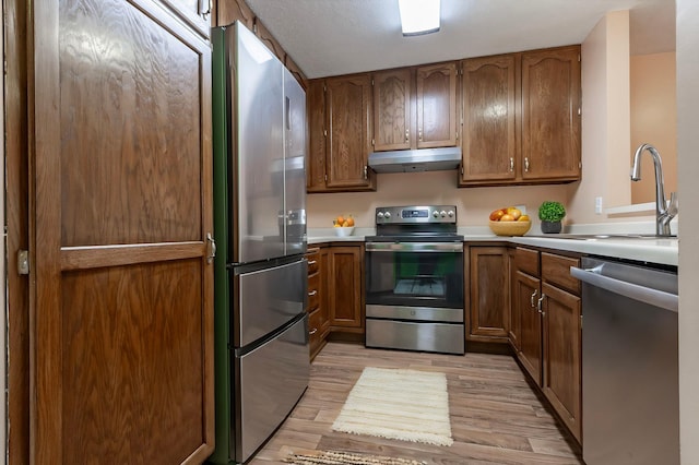 kitchen featuring stainless steel appliances, light wood-style flooring, a sink, brown cabinetry, and under cabinet range hood