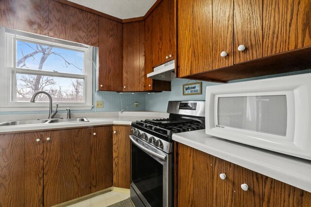 kitchen with a sink, light countertops, under cabinet range hood, stainless steel range with gas stovetop, and white microwave