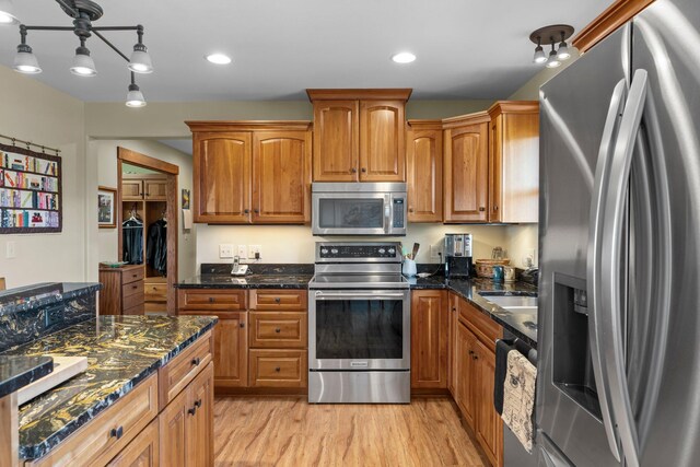 kitchen with brown cabinetry, light wood-type flooring, a sink, dark stone counters, and stainless steel appliances
