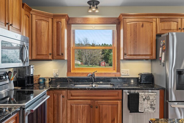 kitchen with a sink, appliances with stainless steel finishes, brown cabinetry, and dark stone countertops