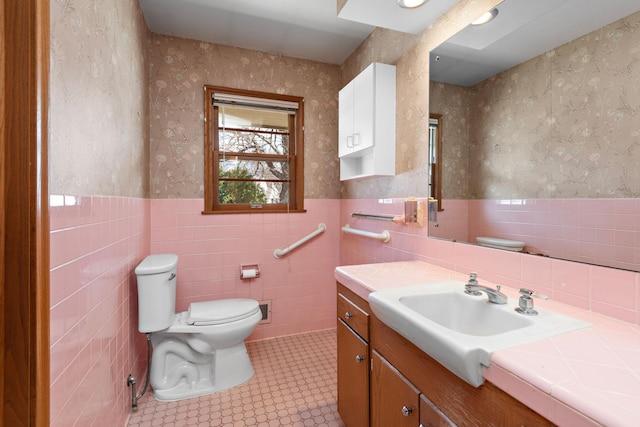 bathroom with toilet, a wainscoted wall, tile walls, vanity, and wallpapered walls