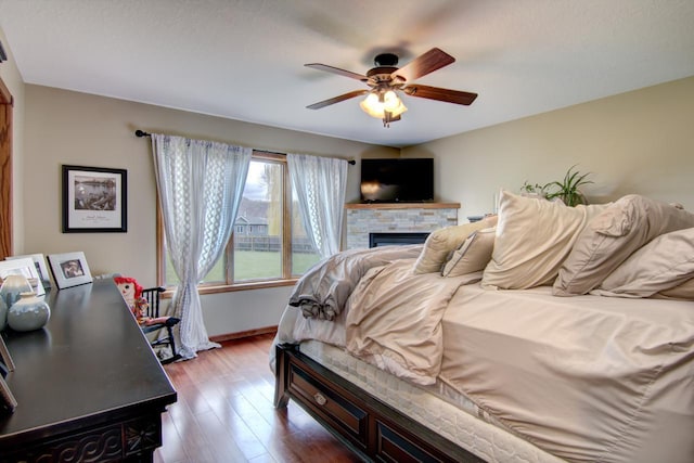 bedroom with baseboards, wood finished floors, a stone fireplace, and a ceiling fan