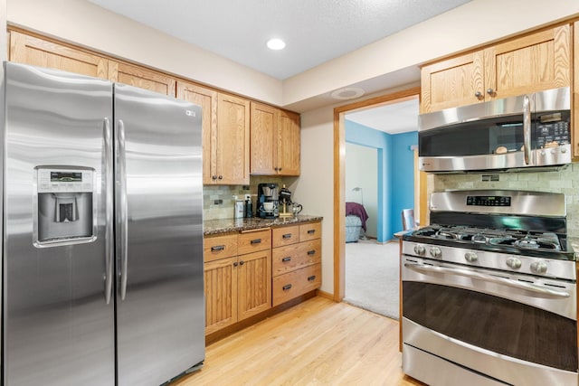 kitchen with stainless steel appliances, light wood-style floors, decorative backsplash, dark stone countertops, and light brown cabinetry