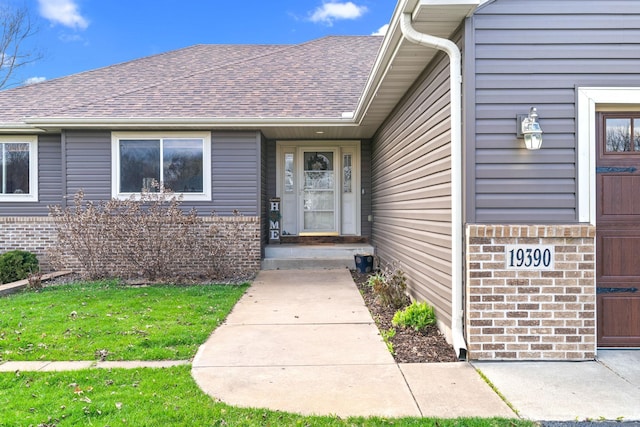doorway to property featuring a shingled roof, brick siding, and a lawn