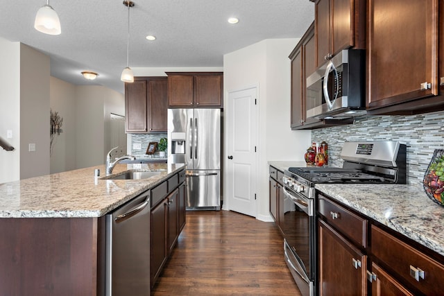 kitchen featuring light stone counters, a sink, an island with sink, stainless steel appliances, and dark wood-style flooring