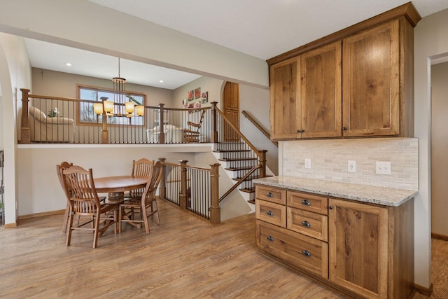 kitchen featuring light wood-type flooring, a chandelier, decorative backsplash, brown cabinets, and light stone countertops