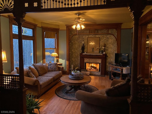 living room with a warm lit fireplace, ornate columns, a ceiling fan, and hardwood / wood-style flooring