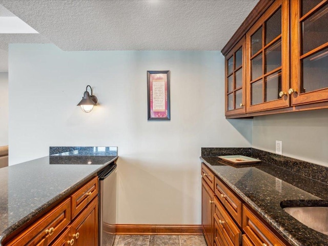 kitchen featuring baseboards, a textured ceiling, brown cabinets, dark stone counters, and glass insert cabinets