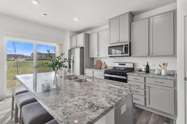kitchen featuring a sink, a kitchen island with sink, gray cabinets, appliances with stainless steel finishes, and light stone countertops