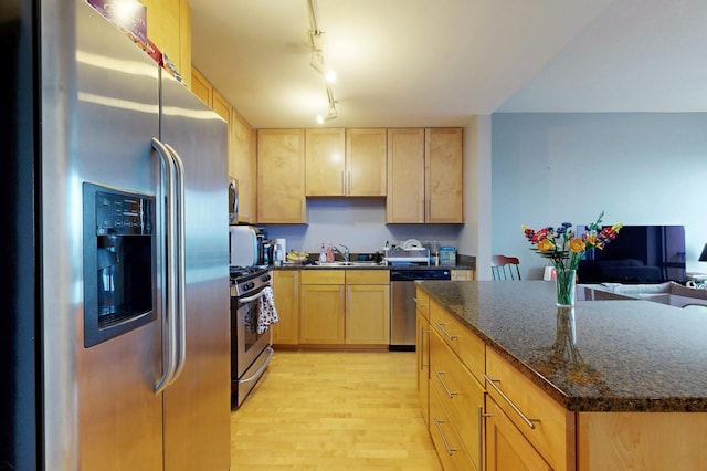 kitchen featuring stainless steel appliances, a sink, light brown cabinets, light wood-style flooring, and dark stone countertops