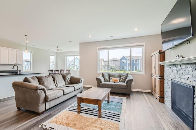 living room featuring a ceiling fan, a stone fireplace, light wood-style flooring, baseboards, and recessed lighting