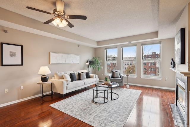 living area featuring a ceiling fan, a textured ceiling, a tiled fireplace, baseboards, and dark wood-style flooring