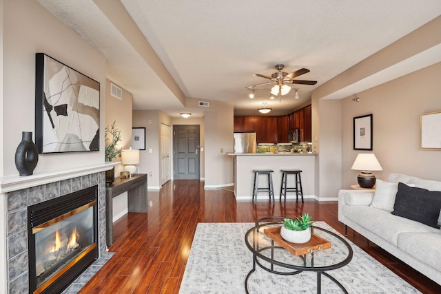living area featuring a tile fireplace, dark wood-style floors, visible vents, a ceiling fan, and baseboards