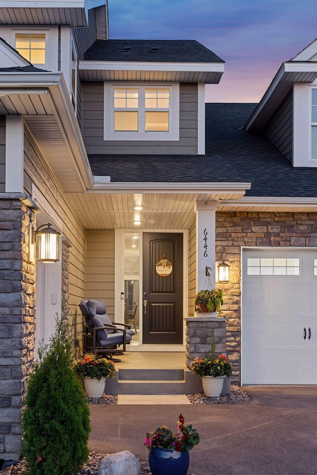 exterior entry at dusk featuring stone siding, covered porch, a shingled roof, driveway, and a garage