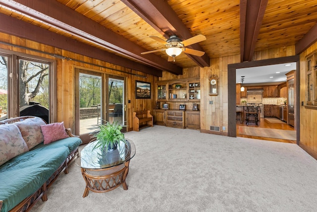 living room featuring wooden walls, beamed ceiling, light colored carpet, a ceiling fan, and wood ceiling