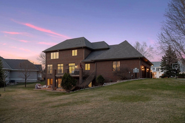 back of property at dusk with a yard, stairs, a shingled roof, and brick siding