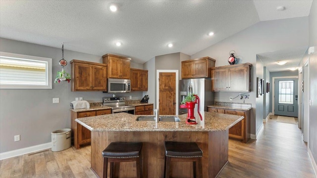 kitchen with light wood-style floors, brown cabinetry, an island with sink, stainless steel appliances, and vaulted ceiling