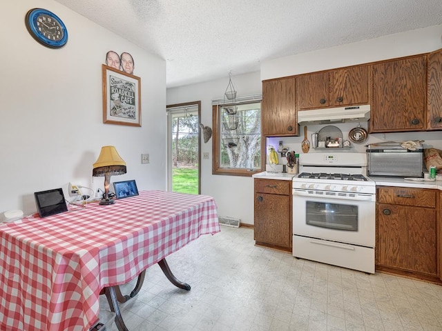 kitchen with brown cabinetry, a textured ceiling, under cabinet range hood, light countertops, and white range with gas stovetop