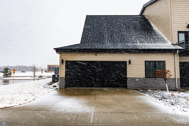 view of snowy exterior with concrete driveway, roof with shingles, and a garage