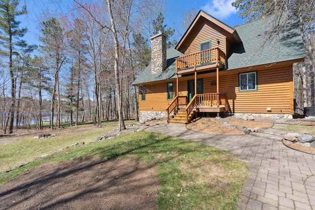 chalet / cabin featuring a balcony, a shingled roof, a chimney, log veneer siding, and a front lawn