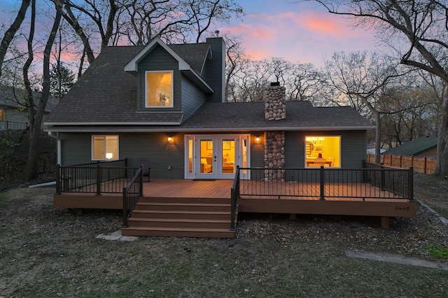 back of house at dusk with roof with shingles, a chimney, french doors, and a wooden deck