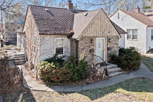 english style home featuring a shingled roof, stucco siding, stone siding, and a chimney