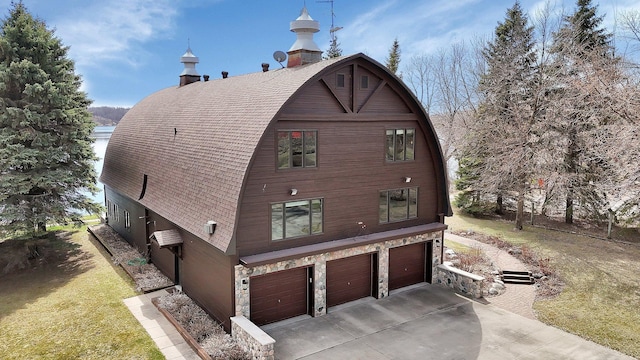 view of property exterior with driveway, stone siding, roof with shingles, an attached garage, and a gambrel roof