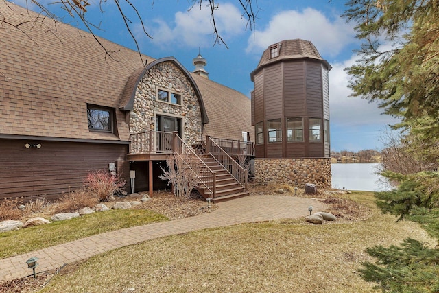 back of property with a deck with water view, stairs, a lawn, stone siding, and roof with shingles