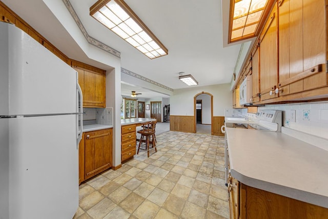 kitchen with brown cabinetry, white appliances, open floor plan, a wainscoted wall, and arched walkways
