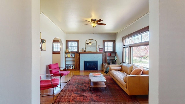 living area with crown molding, arched walkways, a glass covered fireplace, a ceiling fan, and wood finished floors