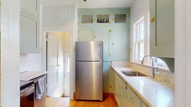 kitchen with light stone countertops, a sink, green cabinets, glass insert cabinets, and stainless steel appliances