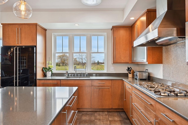 kitchen with a sink, stainless steel gas cooktop, brown cabinetry, wall chimney exhaust hood, and refrigerator with glass door