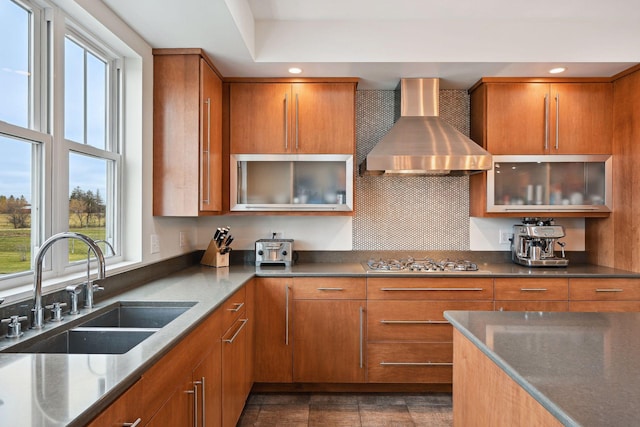 kitchen with glass insert cabinets, brown cabinetry, a sink, stainless steel gas stovetop, and wall chimney exhaust hood