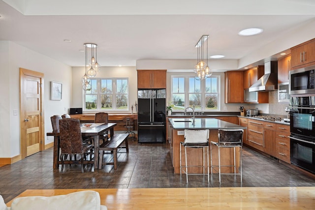 kitchen with a kitchen island with sink, brown cabinetry, dark countertops, black appliances, and wall chimney exhaust hood