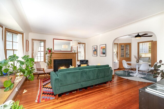 living area featuring wood-type flooring, arched walkways, a lit fireplace, and ceiling fan