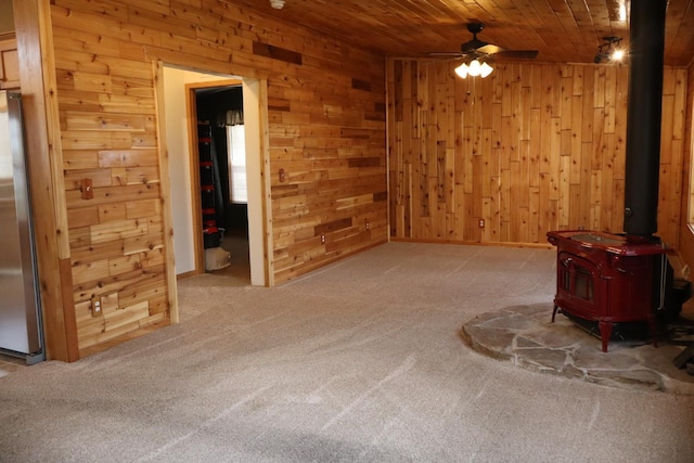 unfurnished living room featuring wooden walls, a wood stove, carpet, wood ceiling, and ceiling fan