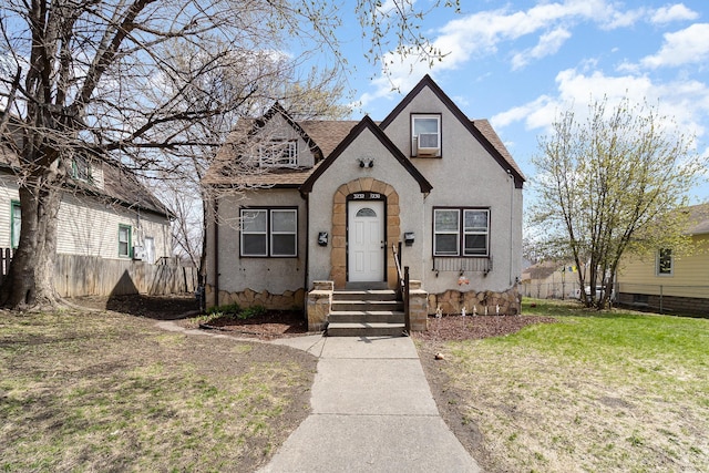 tudor-style house featuring fence, roof with shingles, stucco siding, and a front lawn