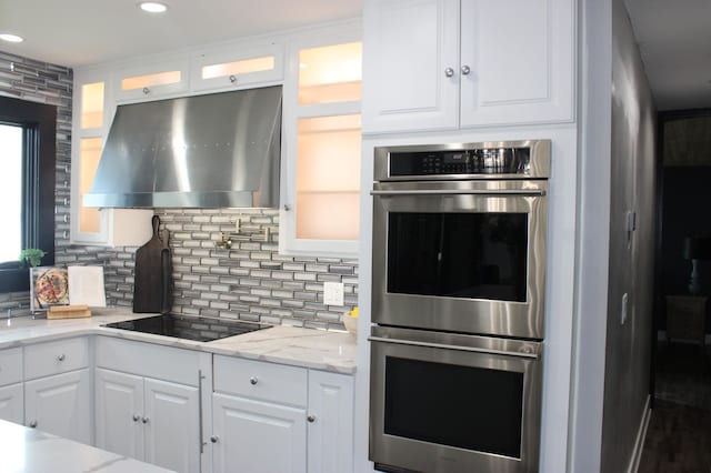 kitchen with stainless steel double oven, range hood, white cabinets, decorative backsplash, and glass insert cabinets
