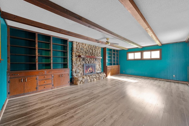 unfurnished living room with wood finished floors, beam ceiling, a textured ceiling, ceiling fan, and a stone fireplace