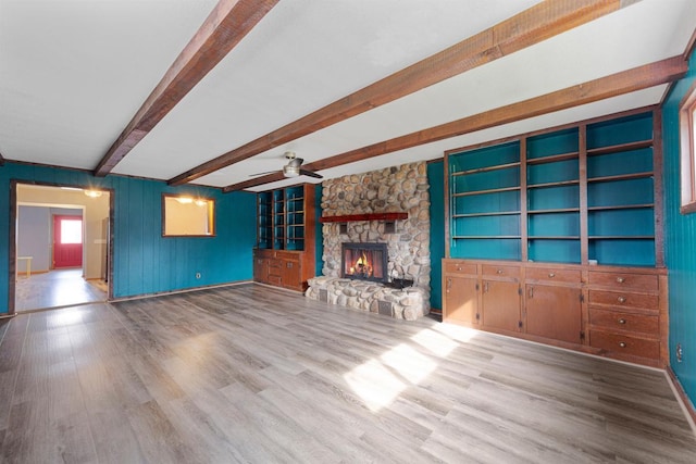 unfurnished living room featuring wood finished floors, beam ceiling, baseboards, a stone fireplace, and ceiling fan
