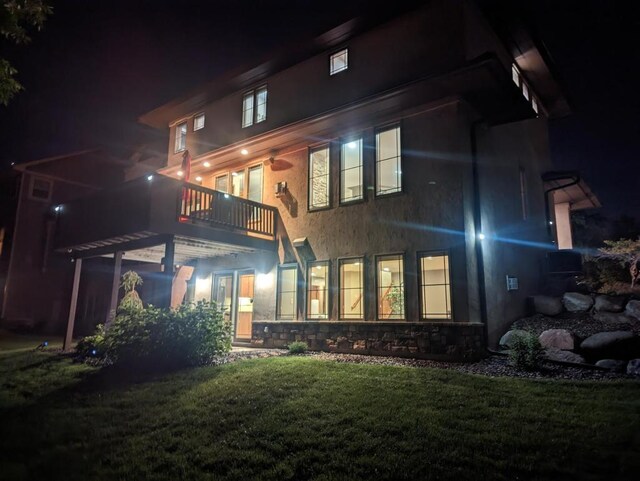 back of house at night with stucco siding, a lawn, stone siding, and a balcony
