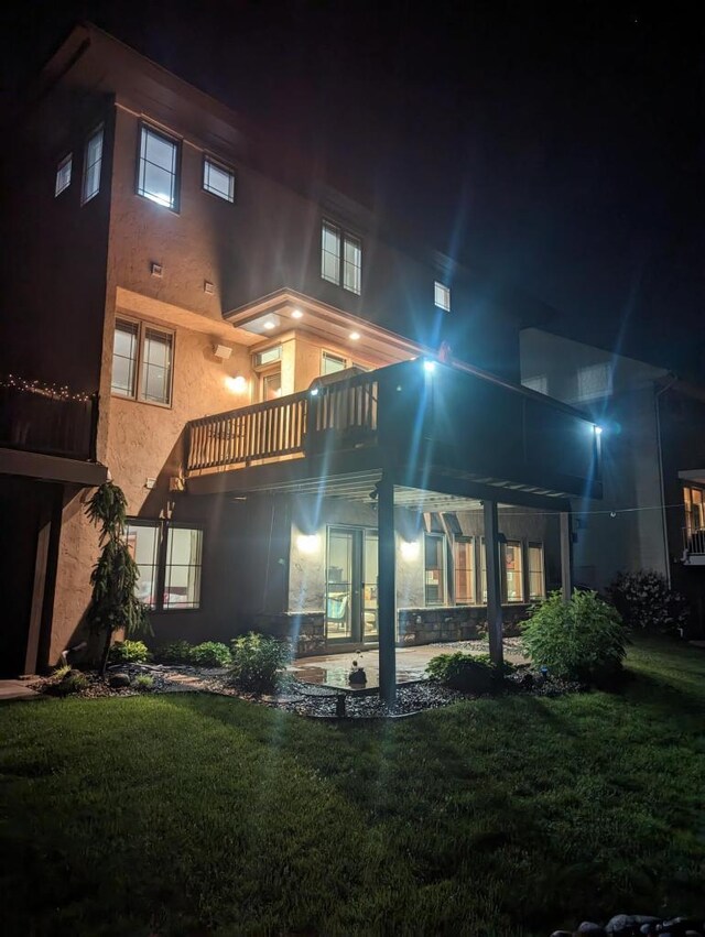 back of house at night featuring stucco siding, a yard, and a balcony