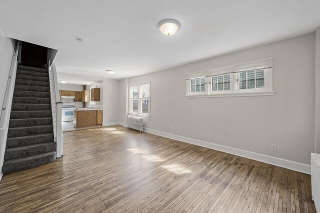 unfurnished living room featuring baseboards, radiator, wood finished floors, and stairway