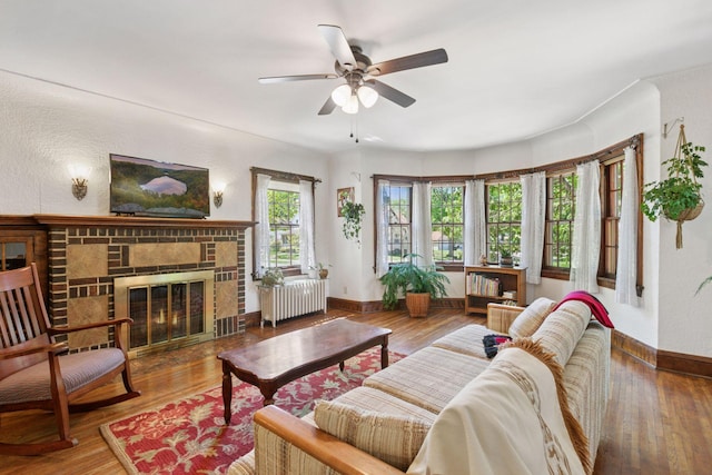 living area featuring radiator, a ceiling fan, baseboards, wood finished floors, and a brick fireplace