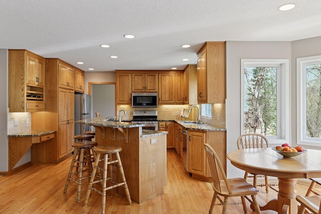 kitchen featuring a sink, appliances with stainless steel finishes, light wood-type flooring, stone counters, and a center island with sink
