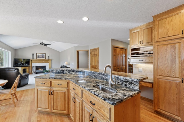 kitchen featuring a sink, light wood-style floors, ceiling fan, a glass covered fireplace, and an island with sink